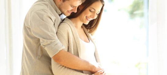 photo of pregnant couple embracing in front of a window babymoon getaway